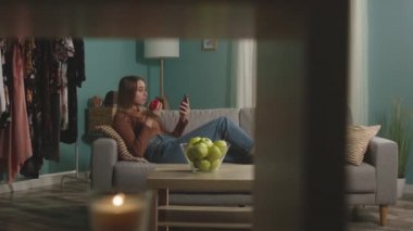 Kırmızı elmalı genç bir kadın kanepede uzanıyor ve bir şeyler izliyor.