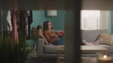 Genç bir kadın kırmızı bir elma yer, kanepede uzanır ve bir şeyler izler.