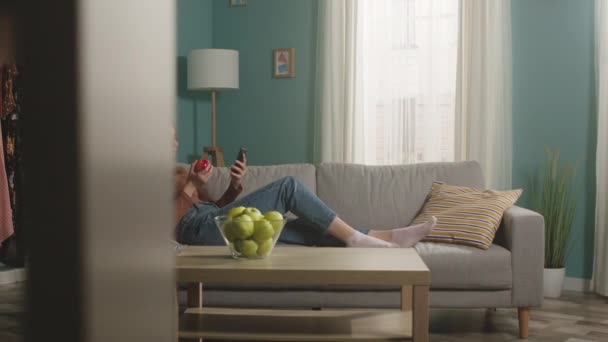 Young woman eats a red apple, lying on sofa and and watching something gadget — Stock Video