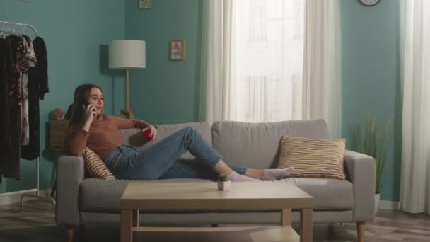 Νεαρή κυρία με κόκκινο μήλο ξαπλώνει στον καναπέ και μιλάει στο τηλέφωνο — Αρχείο Βίντεο