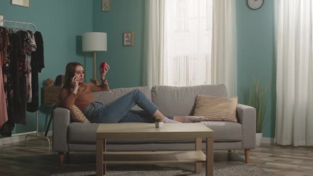 Młoda dziewczyna z czerwonym jabłkiem leży na kanapie i rozmawia przez telefon — Wideo stockowe