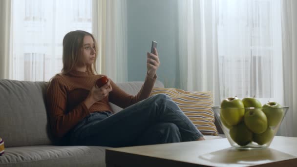 Ung flicka med rött äpple sitter på soffan och tar selfie på mobiltelefon — Stockvideo