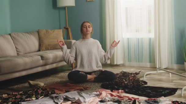 Gyönyörű nő ül a padlón szétszórt ruhák és meditál — Stock videók