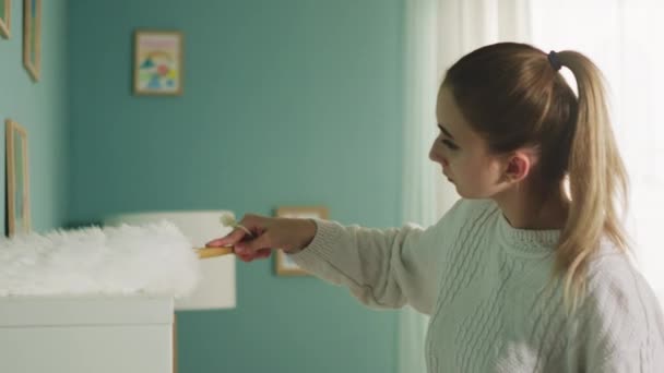 Mladá dívka v bílém svetru otírá prach z nábytku. — Stock video