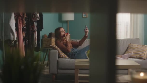 Wanita muda makan apel merah, berbaring di sofa dan mengambil selfie melalui telepon — Stok Video