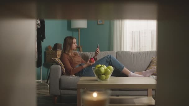 Mulher come uma maçã vermelha, deitada no sofá e fala via link de vídeo — Vídeo de Stock