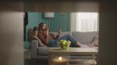 Genç bir kadın kanepede uzanmış kırmızı bir elma yiyor ve video bağlantısıyla konuşuyor.