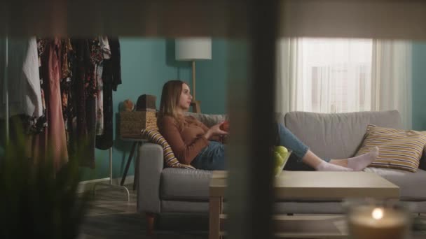 Genç bir kadın kanepede uzanmış kırmızı bir elma yiyor ve video bağlantısıyla konuşuyor. — Stok video