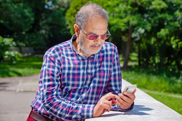 Pensionista serio con gafas de ropa casual y teléfono en el parque — Foto de Stock