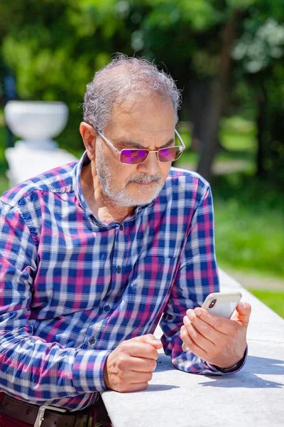 Pensionista serio con gafas de ropa casual y teléfono en el parque — Foto de Stock