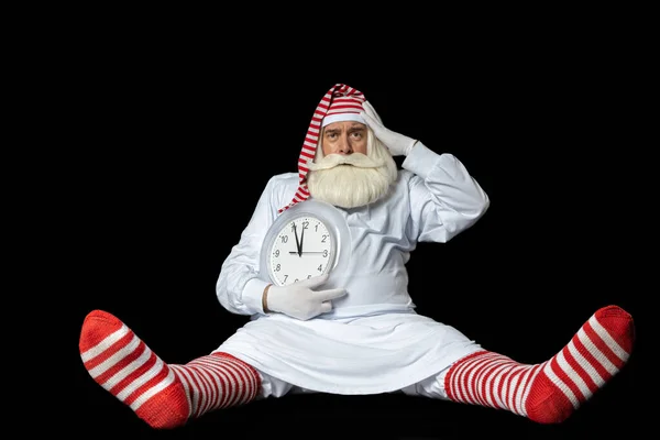 Noel Baba gecelikli ve yatmadan önce elinde saatle yerde oturuyor ve Noel 'i düşünüyor. Siyah bir arkaplanda.