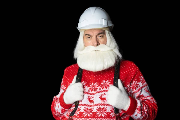 Noel Baba kazaklı, kasklı, gri saçlı bir iş adamı ellerini pantolon askılarından tutup doğrudan kameraya bakıyor. Endüstriyel Noel konsepti.