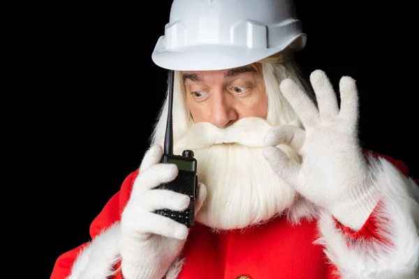 Noel Baba ceketli, miğferli gri saçlı bir adam telsiz kullanıyor ve her şeyin yoluna gireceğini gösteriyor. Siyah soyutlanmış arka plan, Noel endüstrisi konsepti.