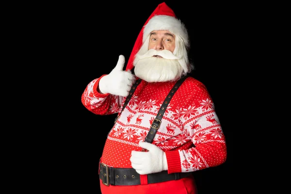 Süveter ve pantolon askısı giymiş yardımsever, sakallı Noel Baba, baş parmağı havada, kopyalama alanında iyi görünüyor. İzole edilmiş siyah arkaplan.