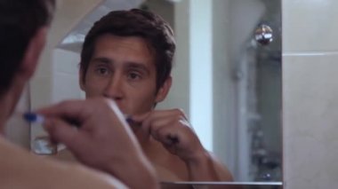 Ciddi bir genç adam banyo aynasının önünde dişlerini fırçalıyor.
