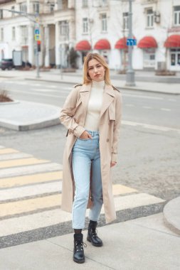 Moda elbiseli güzel sarışın kadın: bej renkli trençkot, siyah çizmeler ve caddeden geçen kot pantolon..