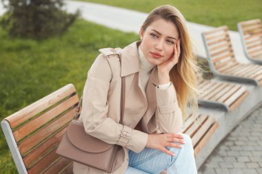 Mavi gözlü, uzun sarı saçlı, sıradan elbiseli, kot ve bej renkli trençkotlu, parkta oturan güzel, üzgün genç bir kadının portresini yakından çek.