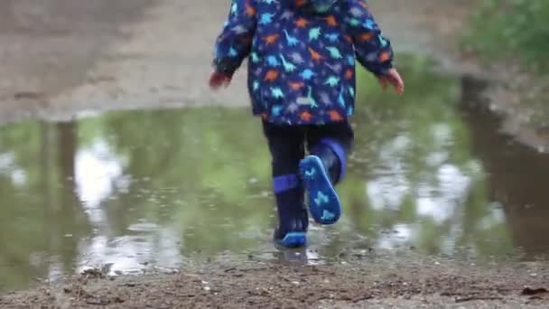 Gelukkig zorgeloos kind loopt door een plas in regenjas en blauwe rubberen laarzen — Stockvideo