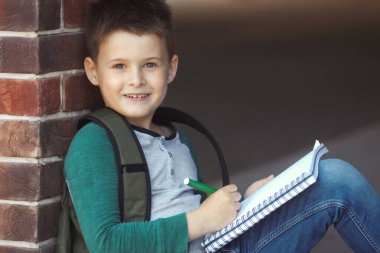 Okul çocuğu, okul bahçesinde duvarın karşısında oturmuş ödevlerini yapıyor ve ebeveynlerini bekliyor.