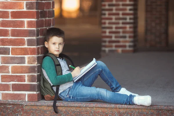 School jongen zit op het schoolplein tegen een bakstenen muur huiswerk te maken en wacht op ouders — Stockfoto