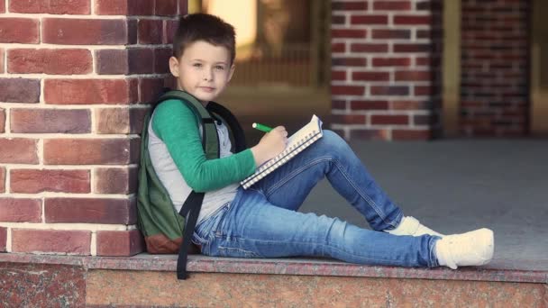 Menino da escola sentado no pátio da escola contra uma parede de tijolo fazendo lição de casa e espera pelos pais — Vídeo de Stock