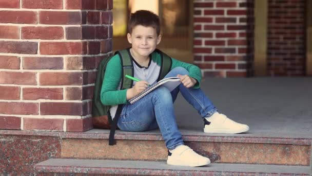 School jongen zit op het schoolplein tegen een bakstenen muur huiswerk te maken en wacht op ouders — Stockvideo