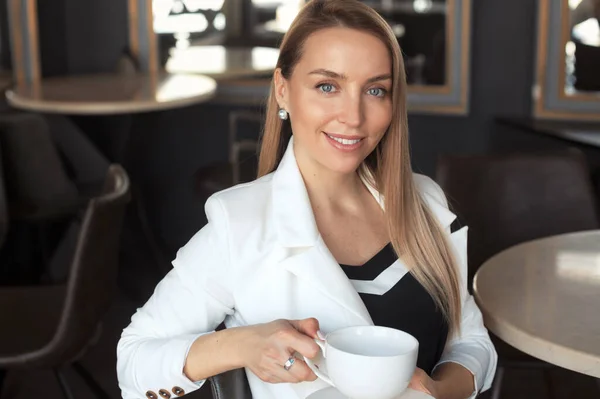 Gülümseyen iş kadınının portresi, sarı saçlı, beyaz ceket giyen ve siyah iç mekânlı kafede kahve içen.