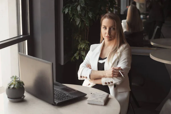 ビジネス女性は白いジャケットでコーヒーを飲み 暗い黒のインテリアのカフェでノートパソコンで作業する — ストック写真