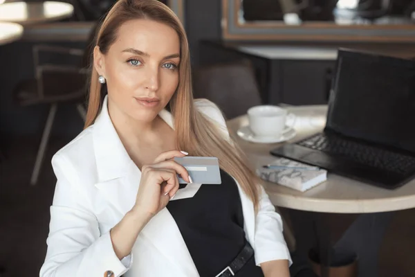 白いブレザーに長いブロンドの髪の飲み物のコーヒーを持つビジネス女性 クレジットカードを保持します — ストック写真