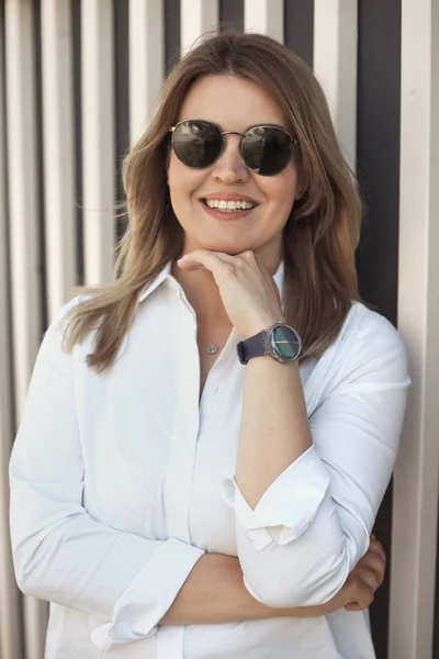 一位30岁至40岁 身穿白衬衫 戴着太阳镜和腕表 面带微笑 自信的年轻女子的生活方式肖像 免版税图库照片