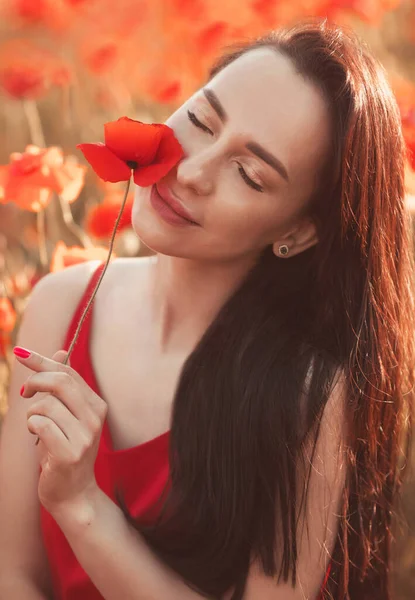 Jovem morena 25-30 anos de idade com cabelos compridos no vestido vermelho desfrutar de flores de papoula no campo — Fotografia de Stock