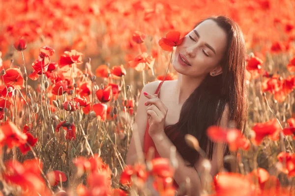 Mujer morena joven de 25-30 años con el pelo largo arreglado en vestido rojo disfrutar de una amapola flores en el campo —  Fotos de Stock