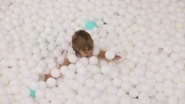 2-3 yaşlarındaki sevimli, oynak çocuk. Beyaz plastik toplarla kuru havuzda eğleniyor.