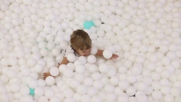 귀엽고 장난기많은 2-3 살 짜리 남자 애가 마른 수영장에서 하얀 플라스틱 공을 가지고 놀고 있어요 — 비디오