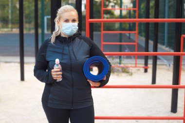 Spor giyim bölümünde yoga minderiyle yorgun, sarışın bir kız sosyal mesafeyi koruyor ve dışarıda antrenman yaparak kilo vermeye çalışıyor. Yaşam tarzı sağlıklı bir kavram. Kilitleme sırasında spor faaliyetleri.