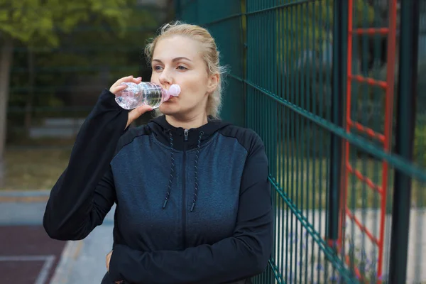 做完运动后 年轻的超重妇女在街头锻炼场所喝水 穿着运动服的疲惫的金发加身材的女孩试图通过户外运动来减肥 高质量的照片 — 图库照片