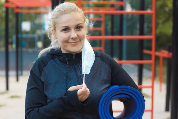 Spor giyim bölümünde yoga minderiyle yorgun, sarışın bir kız sosyal mesafeyi koruyor ve dışarıda antrenman yaparak kilo vermeye çalışıyor. Yaşam tarzı sağlıklı bir kavram. Kilitleme sırasında spor faaliyetleri.