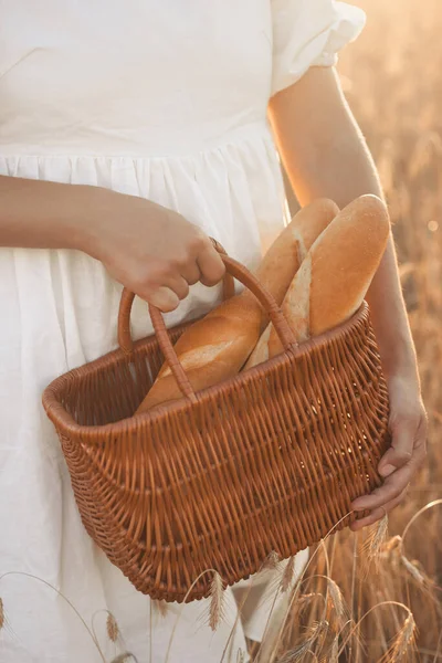 La niña sostiene una cesta de mimbre con pan fresco crujiente al atardecer en el campo de trigo —  Fotos de Stock