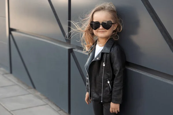 Hezká holčička v módním oblečení: bílé tričko, kožená černá bunda a kamaše, sluneční brýle srdce — Stock fotografie