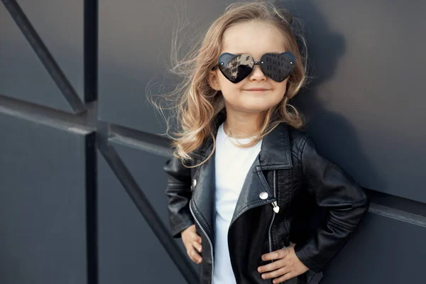 Usmívající se holčička v módním oblečení: bílé tričko, kožená černá bunda a kamaše, sluneční brýle srdce — Stock fotografie