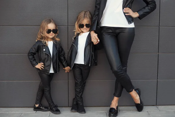 Dua saudara perempuan dengan ibu dalam pakaian yang sama: kaos putih, jaket kulit hitam dan legging, kacamata hitam hati. Stok Gambar