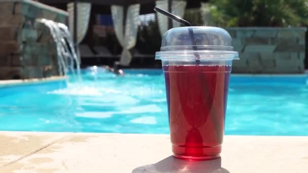 Яскраво-червоний алкогольний коктейль в одноразовій пластиковій чашці з соломою біля басейну — стокове відео