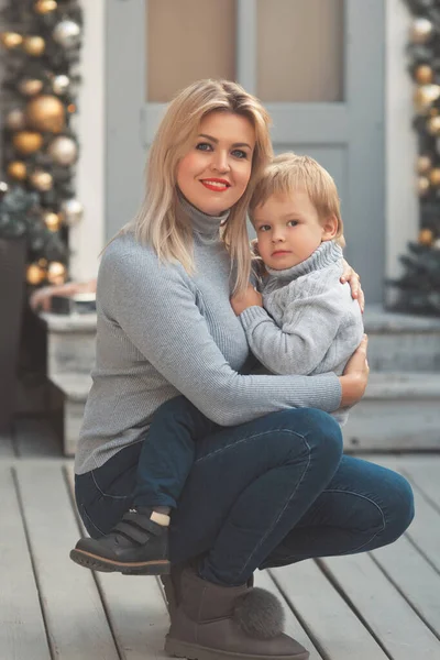 Anne ve çocuk 2-3 yaşlarında, aile görünüşü takınıyorlar. Evlerinin verandasında oturmuş peri masalı gibi Noel süsleriyle.