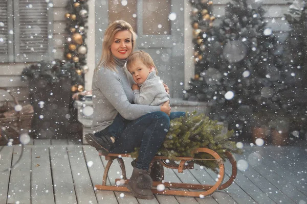 Útulné Vánoce ve sněžném dni. Matka a dítě 2-3 roky sedí na vinobraní dřevěné sáně v novoroční výzdobě v blízkosti verandy domu — Stock fotografie