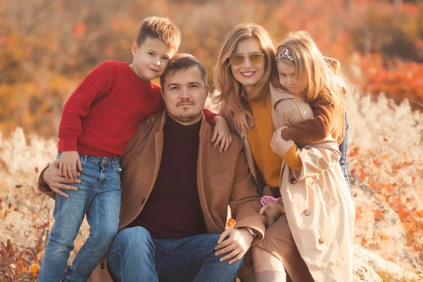 Happy family hugging on a walk in the fall park. Portrait of a caucasian mother and father holding their children in beautiful outfits on a sunny autumn day in forest. Family lifestyle concept.