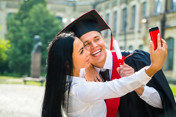 Male graduate and girl taking selfe with cellphone outside — Stock Photo, Image