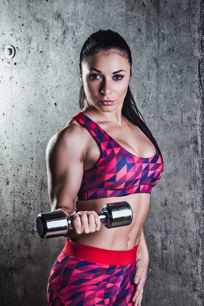 Bella donna muscolosa che fa esercizio con manubri su uno sfondo grigio. Parete di cemento sullo sfondo — Foto Stock