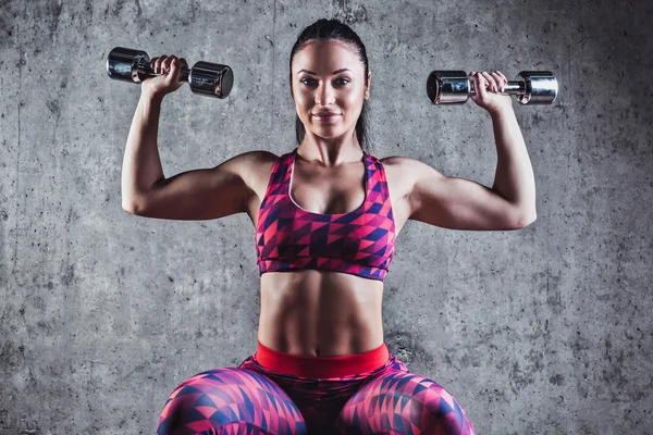Mulher muscular bonita fazendo exercício com halteres em um fundo cinzento.Parede de concreto no fundo — Fotografia de Stock