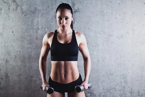 Bella donna muscolosa che fa esercizio con manubri su uno sfondo grigio . — Foto Stock