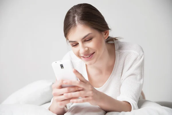 Morena sonriendo mientras usa el teléfono inteligente en la almohada en casa —  Fotos de Stock